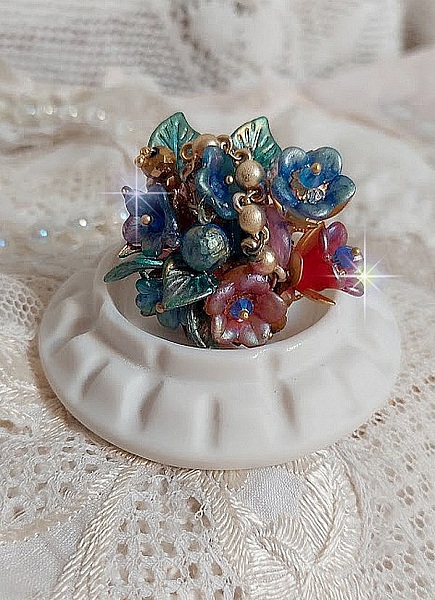 Bague Bel Air mont&eacute;e avec des fleurs lucites peintes &agrave; la main aux couleurs : Bleu, Rose, Rouge, Argent, Blanc avec feuilles ondul&eacute;es Vert Cuivr&eacute; &agrave; laquelle s&rsquo;ajoute perles Dor&eacute;es en Verre