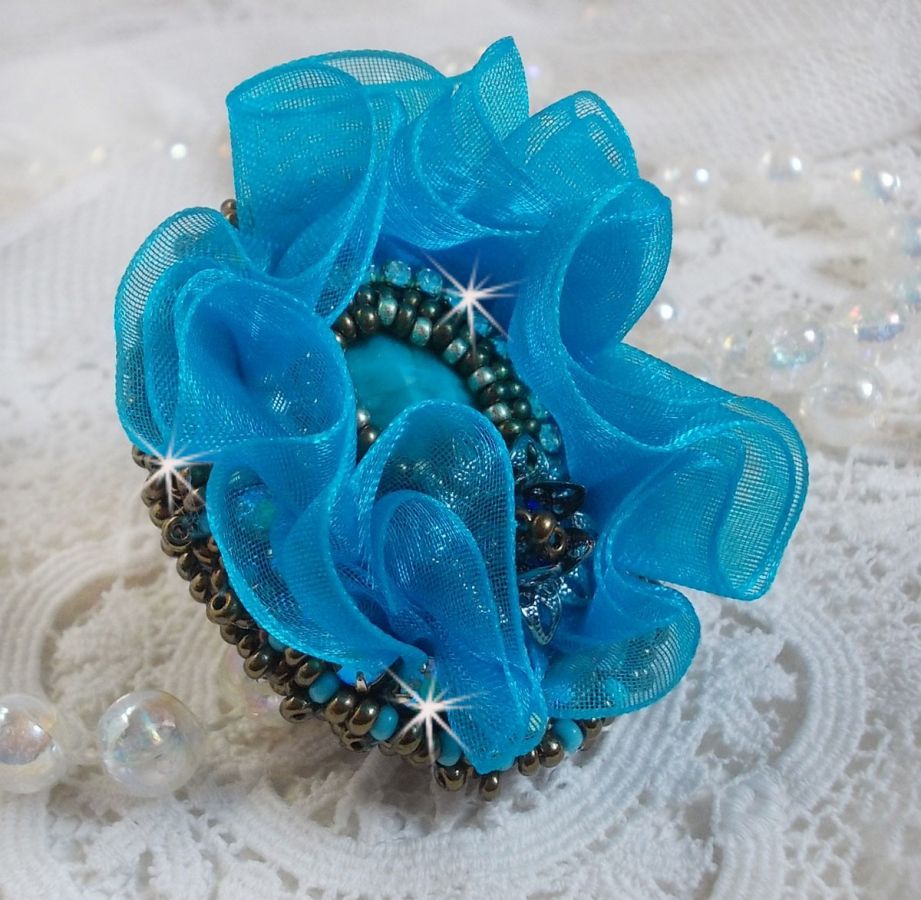 Ilycia Charmig ring broderad med en facetterad turkosblå harts cabochon och ett organza band.