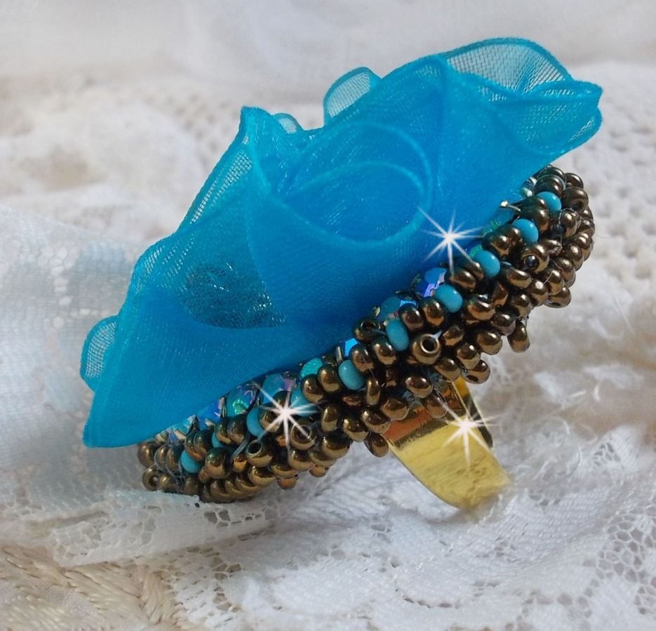 Ilycia Charmig ring broderad med en facetterad turkosblå harts cabochon och ett organza band.