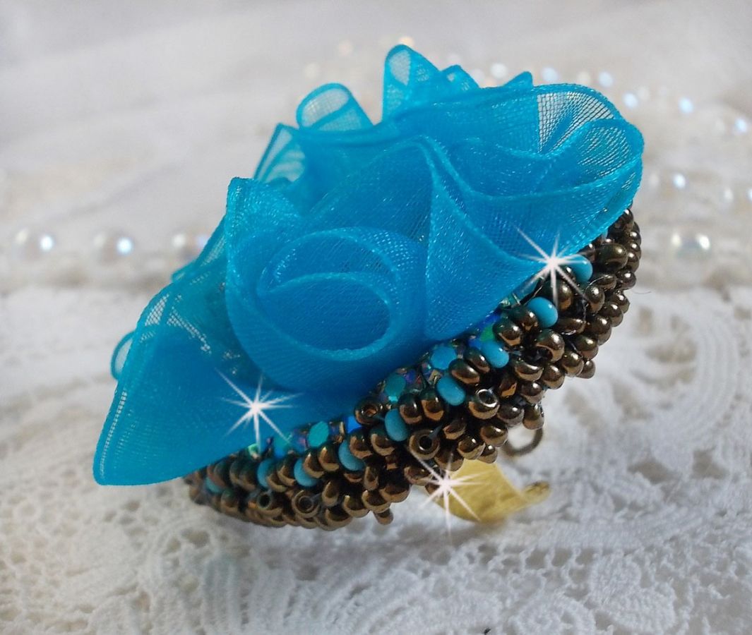 Ilycia Charmig ring broderad med en facetterad turkosblå harts cabochon och ett organza band.