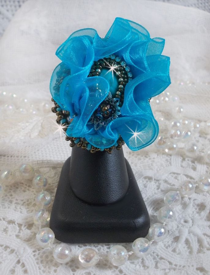 Ilycia Charmig ring broderad med en facetterad turkosblå harts cabochon och ett organza band.