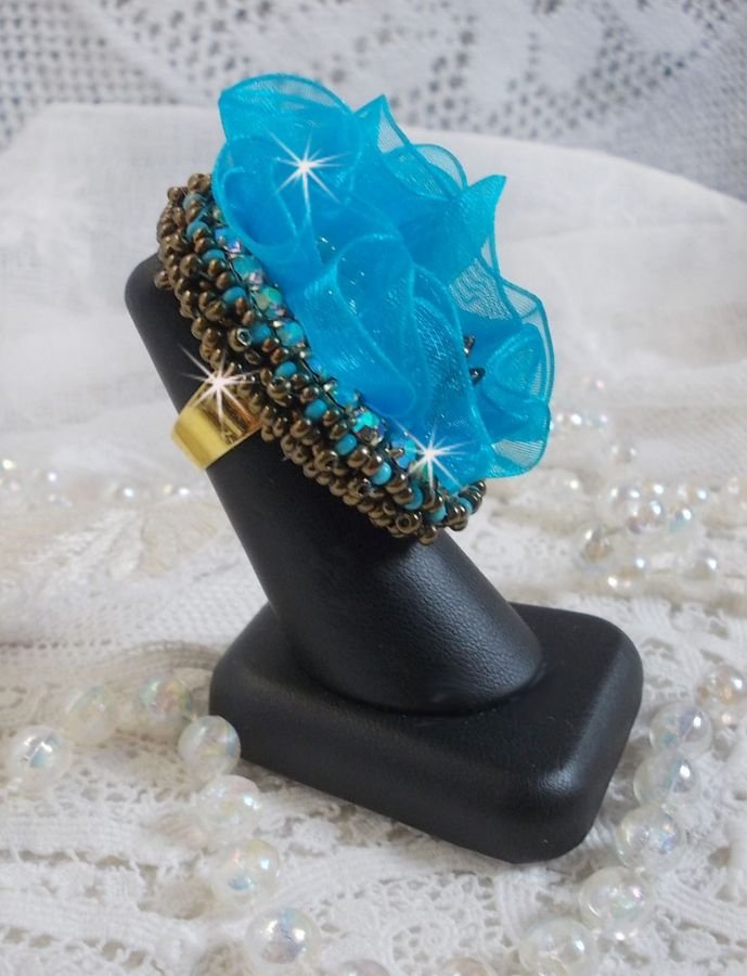 Ilycia Charmig ring broderad med en facetterad turkosblå harts cabochon och ett organza band.