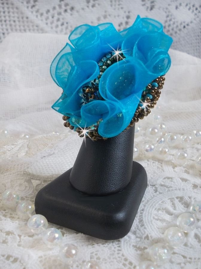 Ilycia Charmig ring broderad med en facetterad turkosblå harts cabochon och ett organza band.