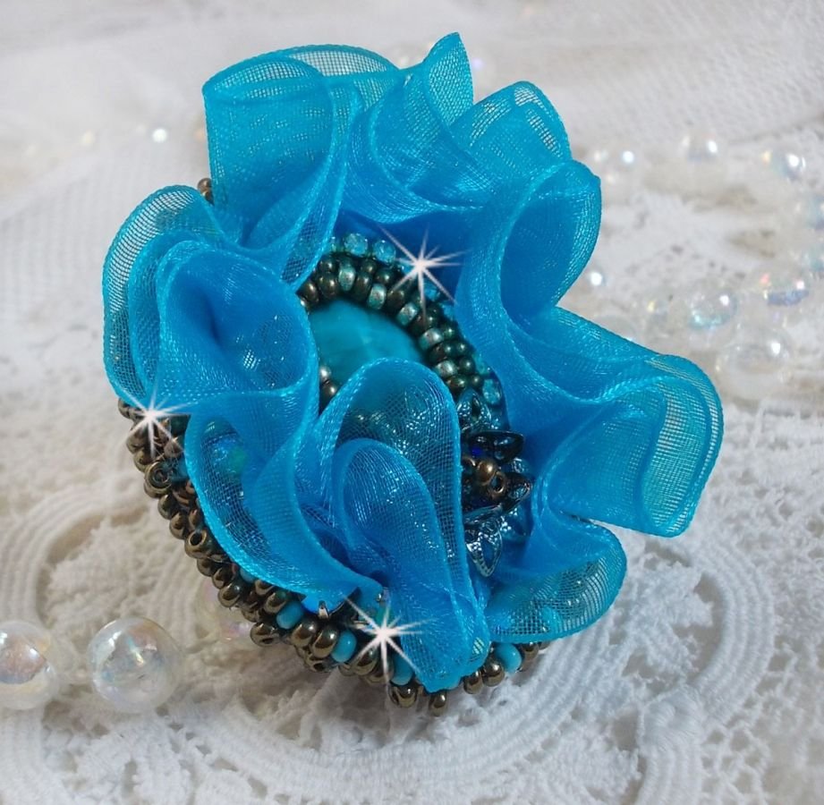 Ilycia Charmig ring broderad med en facetterad turkosblå harts cabochon och ett organza band.