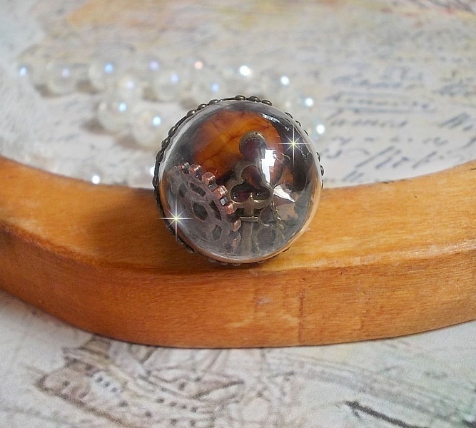 Bague Majest&eacute; Steampunk aux rouages m&eacute;caniques &agrave; laquelle s&rsquo;ajoute une cl&eacute; et un strass de couleur Siam et un strass en verre Dor&eacute;. Le tout est mis sous un globe en verre sur une bague r&eacute;glable laiton