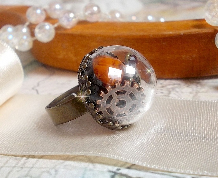 Bague Majest&eacute; Steampunk aux rouages m&eacute;caniques &agrave; laquelle s&rsquo;ajoute une cl&eacute; et un strass de couleur Siam et un strass en verre Dor&eacute;. Le tout est mis sous un globe en verre sur une bague r&eacute;glable laiton