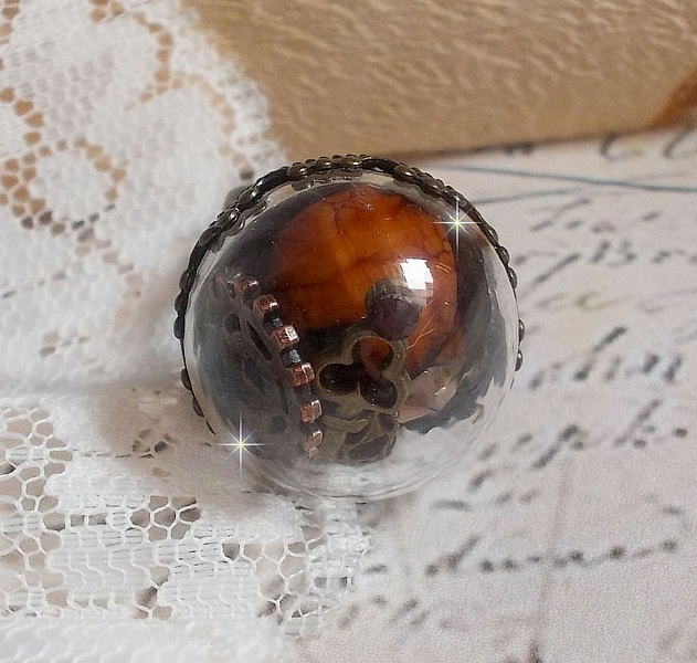 Bague Majest&eacute; Steampunk aux rouages m&eacute;caniques &agrave; laquelle s&rsquo;ajoute une cl&eacute; et un strass de couleur Siam et un strass en verre Dor&eacute;. Le tout est mis sous un globe en verre sur une bague r&eacute;glable laiton