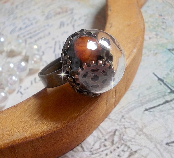 Bague Majest&eacute; Steampunk aux rouages m&eacute;caniques &agrave; laquelle s&rsquo;ajoute une cl&eacute; et un strass de couleur Siam et un strass en verre Dor&eacute;. Le tout est mis sous un globe en verre sur une bague r&eacute;glable laiton