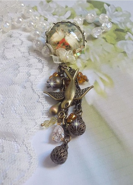 Broche Colombes aux plumages blancs avec des breloques filigran&eacute;es gouttes et lisses, perles rondes et n&oelig;ud en cristal, ruban organza cr&egrave;me, pendentif plaqu&eacute; or avec un zircon sur une broche en m&eacute;tal