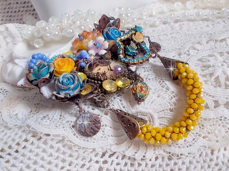 Broche Eclats P&eacute;tillants d&rsquo;inspiration Baroque, orn&eacute;e de fleurs en r&eacute;sine autour d&rsquo;un cabochon strass&eacute;s et perl&eacute;s sur estampes, cristaux, breloques, perle cloisonn&eacute;e, perles jaunes en verre et roses. 