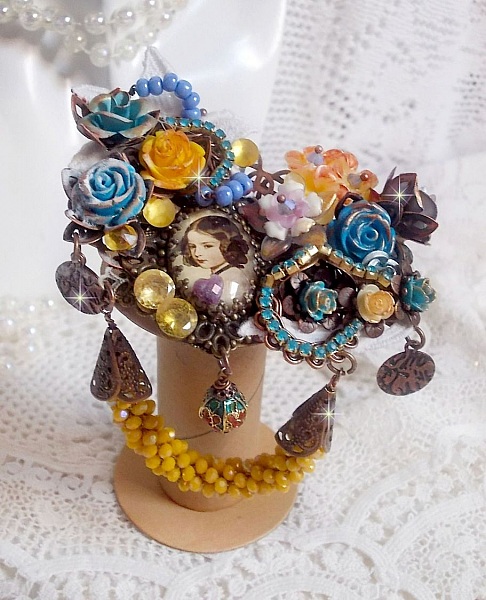 Broche Eclats P&eacute;tillants d&rsquo;inspiration Baroque, orn&eacute;e de fleurs en r&eacute;sine autour d&rsquo;un cabochon strass&eacute;s et perl&eacute;s sur estampes, cristaux, breloques, perle cloisonn&eacute;e, perles jaunes en verre et roses. 